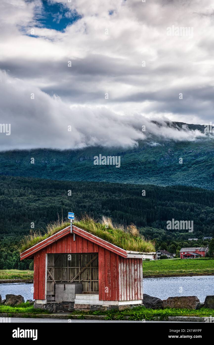 Il tradizionale rifugio rosso degli autobus con un tetto verde si erge davanti a una spettacolare e montuosa nuvole di cumulus sullo sfondo nel villaggio di Halsa, Trøndelag, Norvegia Foto Stock
