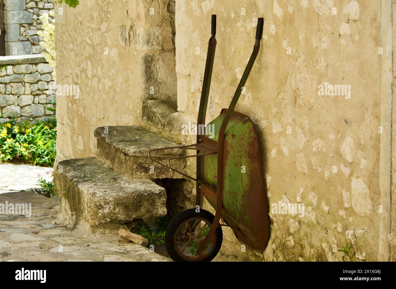 La vecchia carriola arrugginita comune è posizionata contro un edificio in pietra di fronte a una scala in Francia. Foto Stock