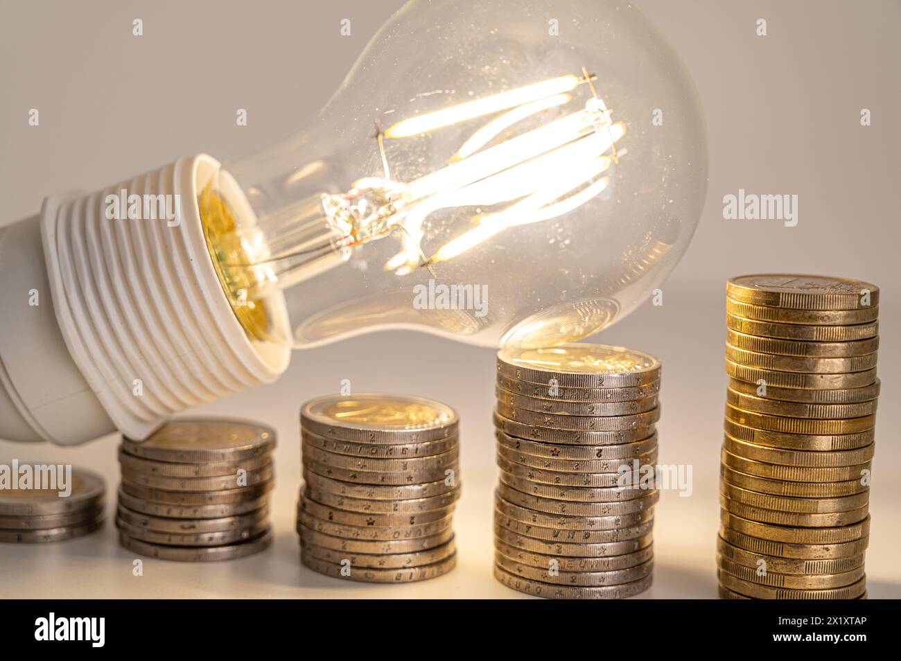 Lampadina accesa, sopra pile di monete. Aumento delle tariffe elettriche, della dipendenza energetica, delle forniture energetiche. Foto Stock