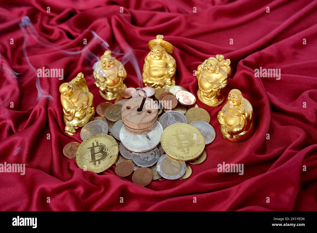 Scivolo di monete metalliche, valute di diversi paesi, bitcoin su tendaggi rossi di lusso, cinque hotei giapponesi dio della ricchezza, un bastone di incenso è bruciato Foto Stock