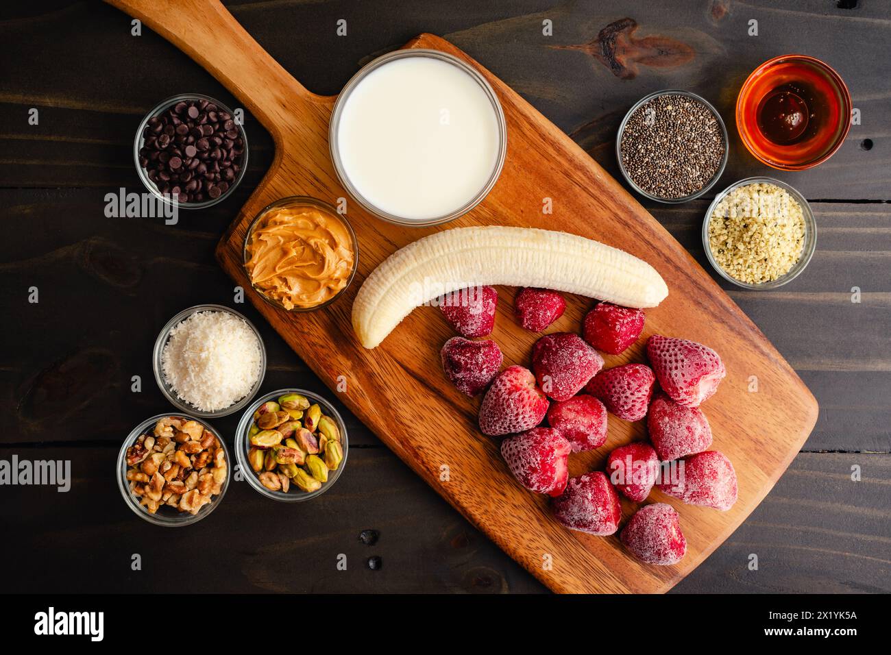 Ingredienti della ciotola per frullati alla fragola su un tagliere di legno: Fragole surgelate e banana con latte e vari condimenti morbidi Foto Stock