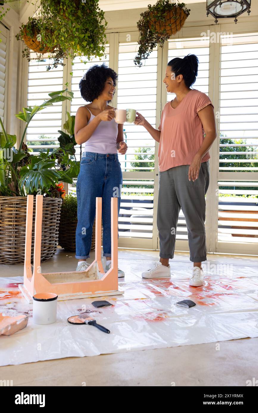 Madre birazziale e figlia adulta che si gusta un caffè durante il progetto di pittura a casa e upcycling a casa. Entrambi hanno capelli neri ricci, adulti daughte Foto Stock