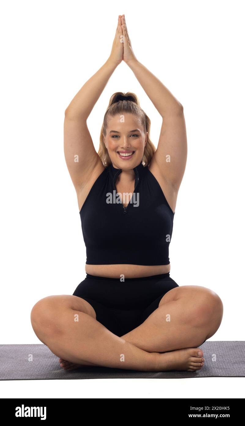 Giovane donna caucasica modello Plus size su sfondo bianco che pratica yoga, seduto sul tappetino. Ha i capelli biondi, gli occhi blu, indossa abiti sportivi neri, Foto Stock