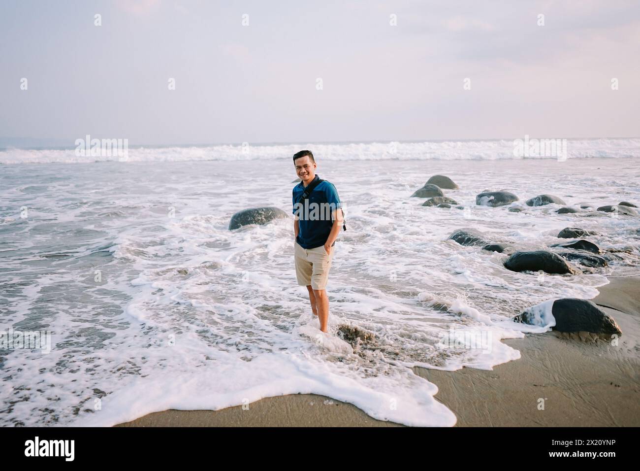 Un uomo soddisfatto con una maglietta blu e pantaloncini si trova sulla riva, il suo sguardo fisso verso l'orizzonte, irradiando felicità sullo sfondo del beac Foto Stock