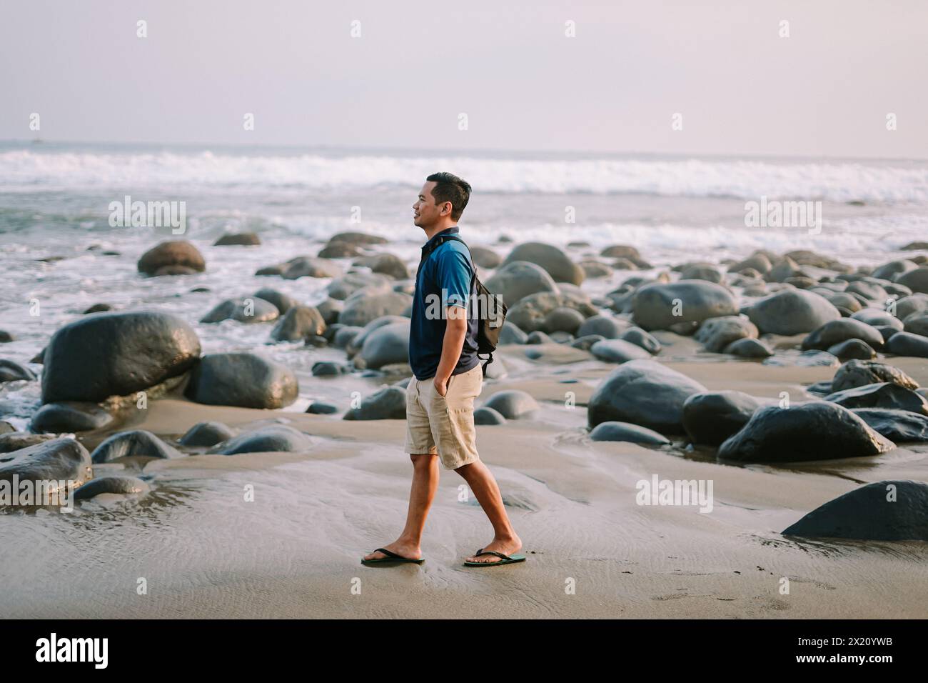 Un uomo soddisfatto con una maglietta blu e pantaloncini si trova sulla riva, il suo sguardo fisso verso l'orizzonte, irradiando felicità sullo sfondo del beac Foto Stock