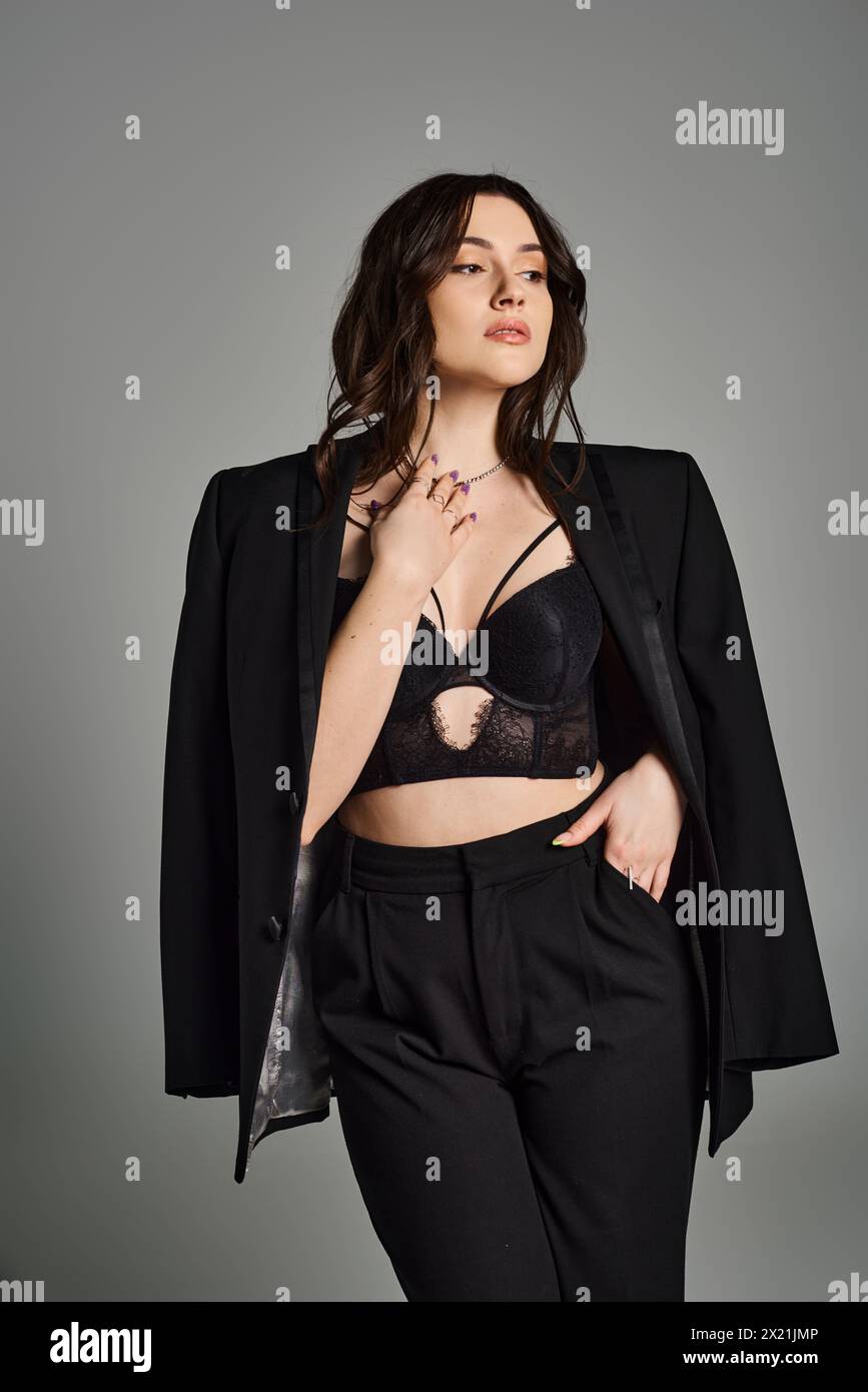 Una bella donna taglia Plus che indossa con sicurezza un elegante bra top nero e pantaloni su uno sfondo grigio. Foto Stock