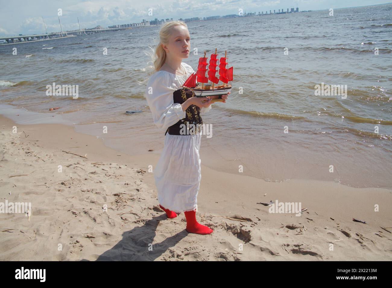 Romantica giovane donna adulta che cammina e tiene in mano un piccolo yacht con vele rosse sulla spiaggia di mare Foto Stock