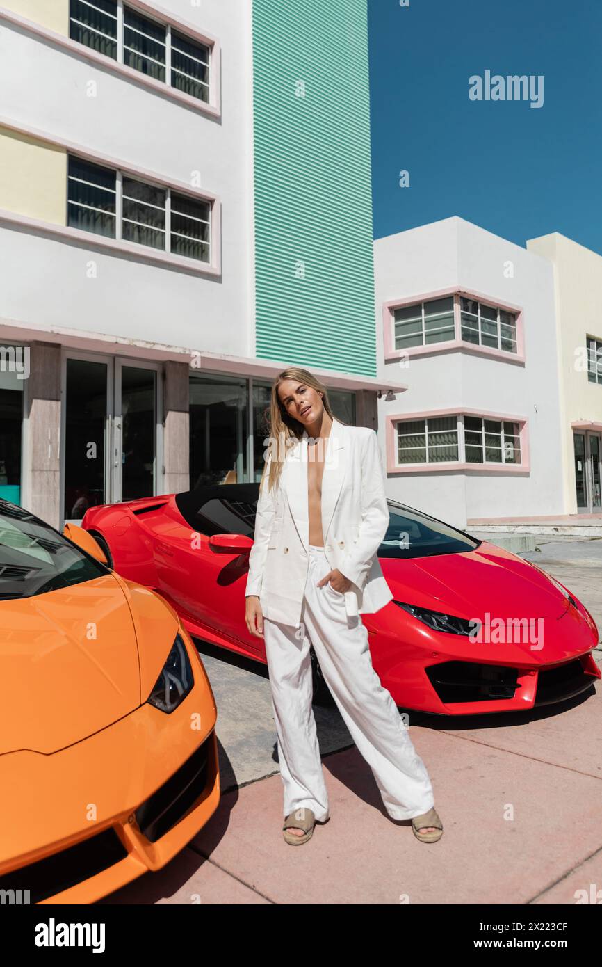 Una giovane e bella donna bionda si erge con fiducia accanto a due eleganti auto sportive a Miami. Foto Stock