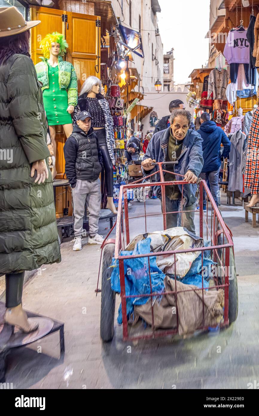 Un uomo in una vivace zona di mercato spinge un carro pieno di merci come parte del trasporto tradizionale non motorizzato nella Medina di Fez, in Marocco. Foto Stock