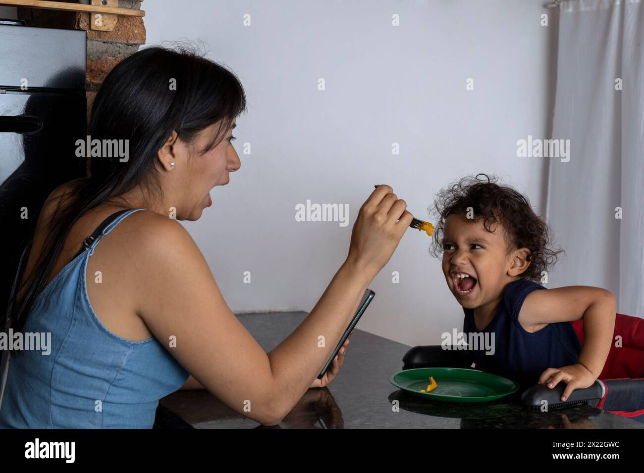 Il ragazzo latino-americano seduto sulla sedia ama essere nutrito dalla madre, fare gesti felici e divertirsi. Concetto di maternità e felicità Foto Stock