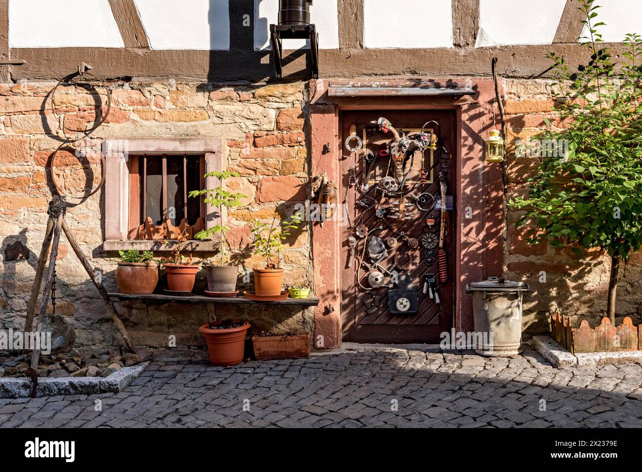 Vecchia casa in legno, decorata in modo stravagante, treppiede con pendolo, vasi da fiori, porta con maschere, teschi, parti del motore, custodie per cartucce, attrezzi, vecchi Foto Stock