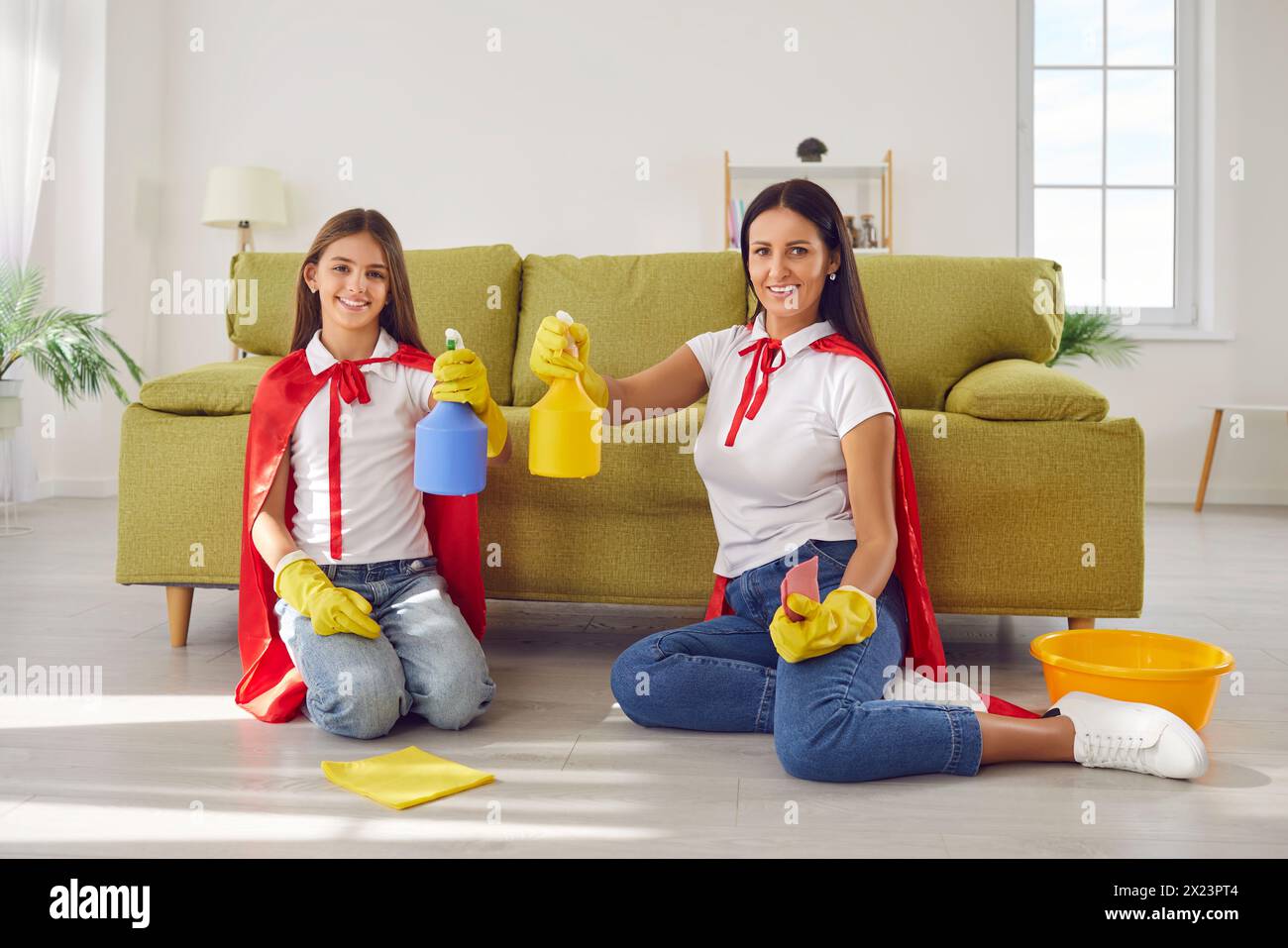 Felice madre sorridente con figlia in costumi da supereroi che puliscono il pavimento a casa. Foto Stock