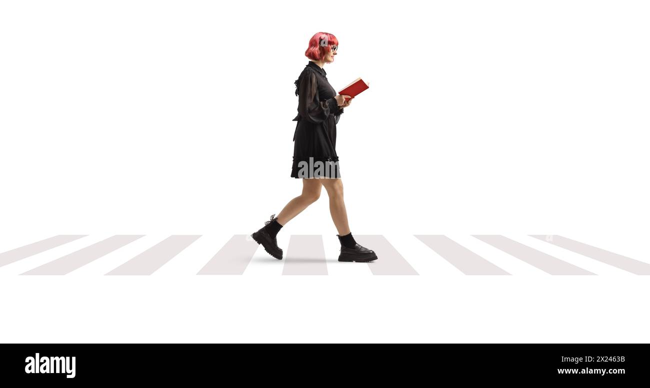 Donna con capelli rossi e abito nero che cammina e legge un libro in un attraversamento pedonale isolato su sfondo bianco Foto Stock