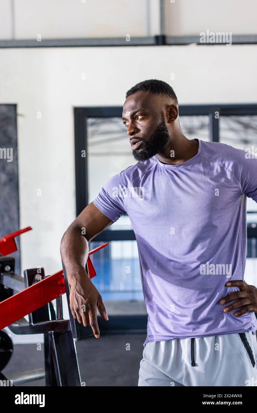 Giovane atleta afroamericano che si appoggia alle attrezzature da palestra in palestra, facendo una pausa Foto Stock