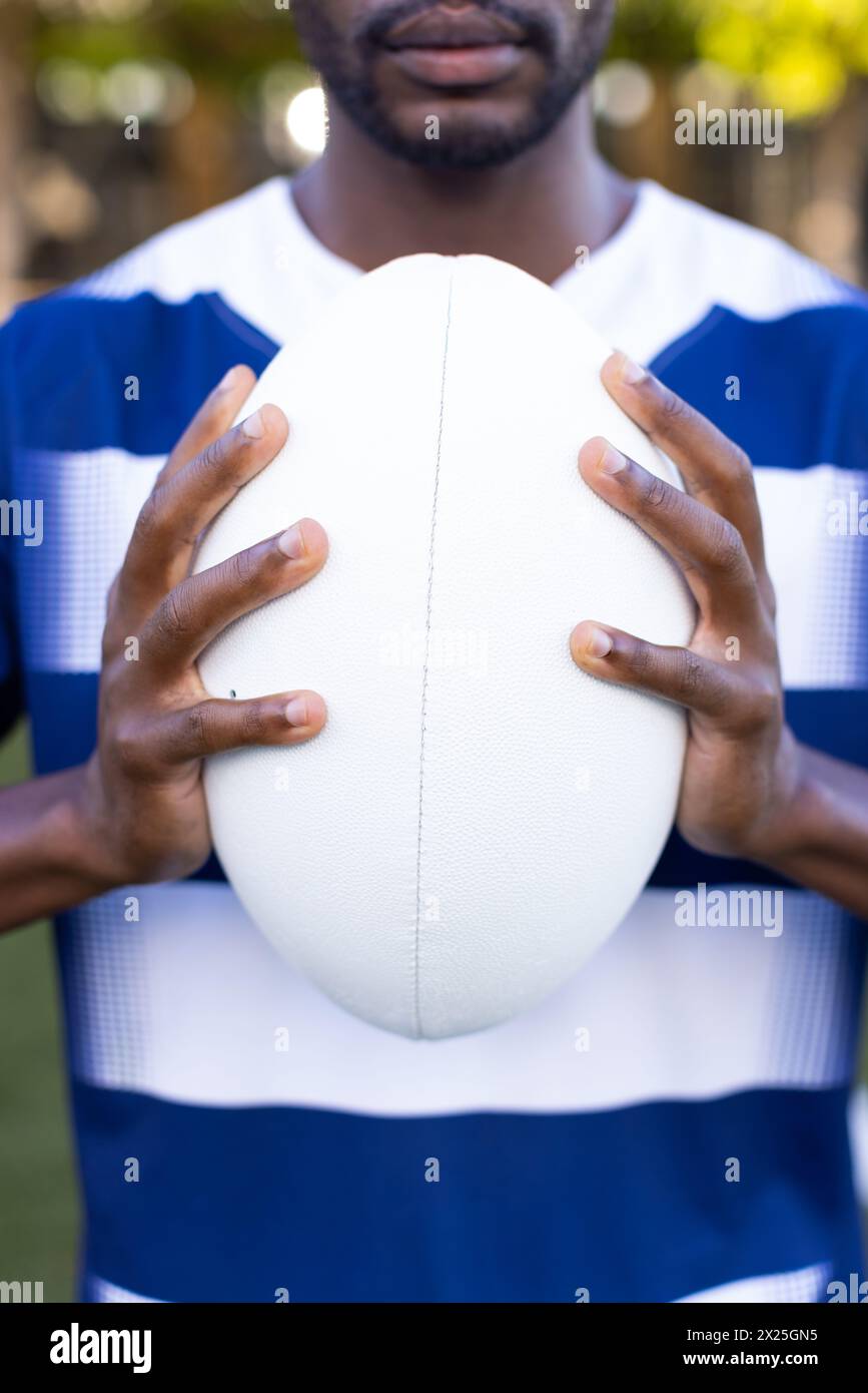 Giovane atleta afroamericano sul campo all'aperto che tiene una palla da rugby e indossa una camicia a righe Foto Stock