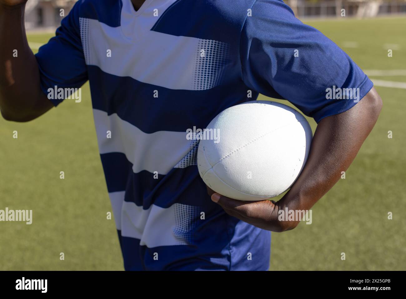 Giovane atleta afroamericano in maglia sportiva a righe che tiene la palla da rugby sul campo all'aperto Foto Stock