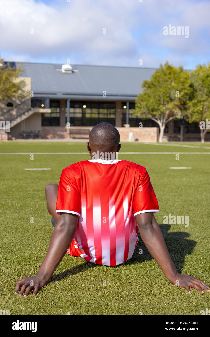 Giovane atleta afroamericano seduto sull'erba sul campo all'aperto, rivolto verso l'esterno Foto Stock