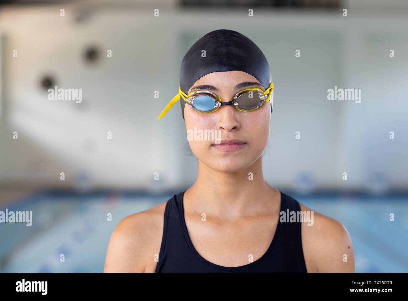 Giovane nuotatrice birazziale in piedi all'interno di fronte a una piscina, indossando occhiali di protezione Foto Stock