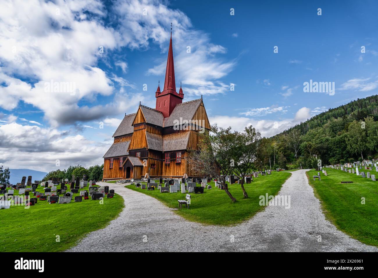 La vista della storica chiesa di Ringebu Stave si erge tra lapidi sotto un cielo blu con nuvole in estate, che riecheggia secoli di storia norvegese. Foto Stock