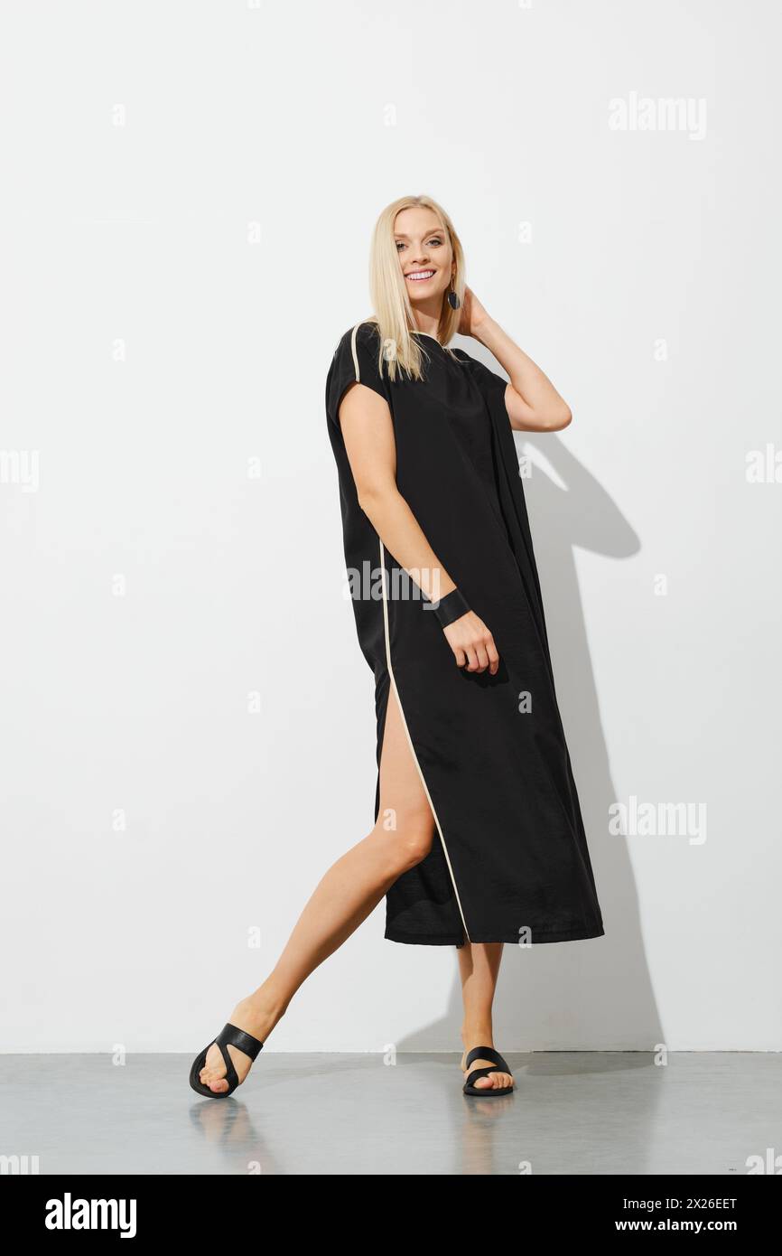 Bella bionda in abito senza maniche nero che posa accanto alla parete bianca Foto Stock