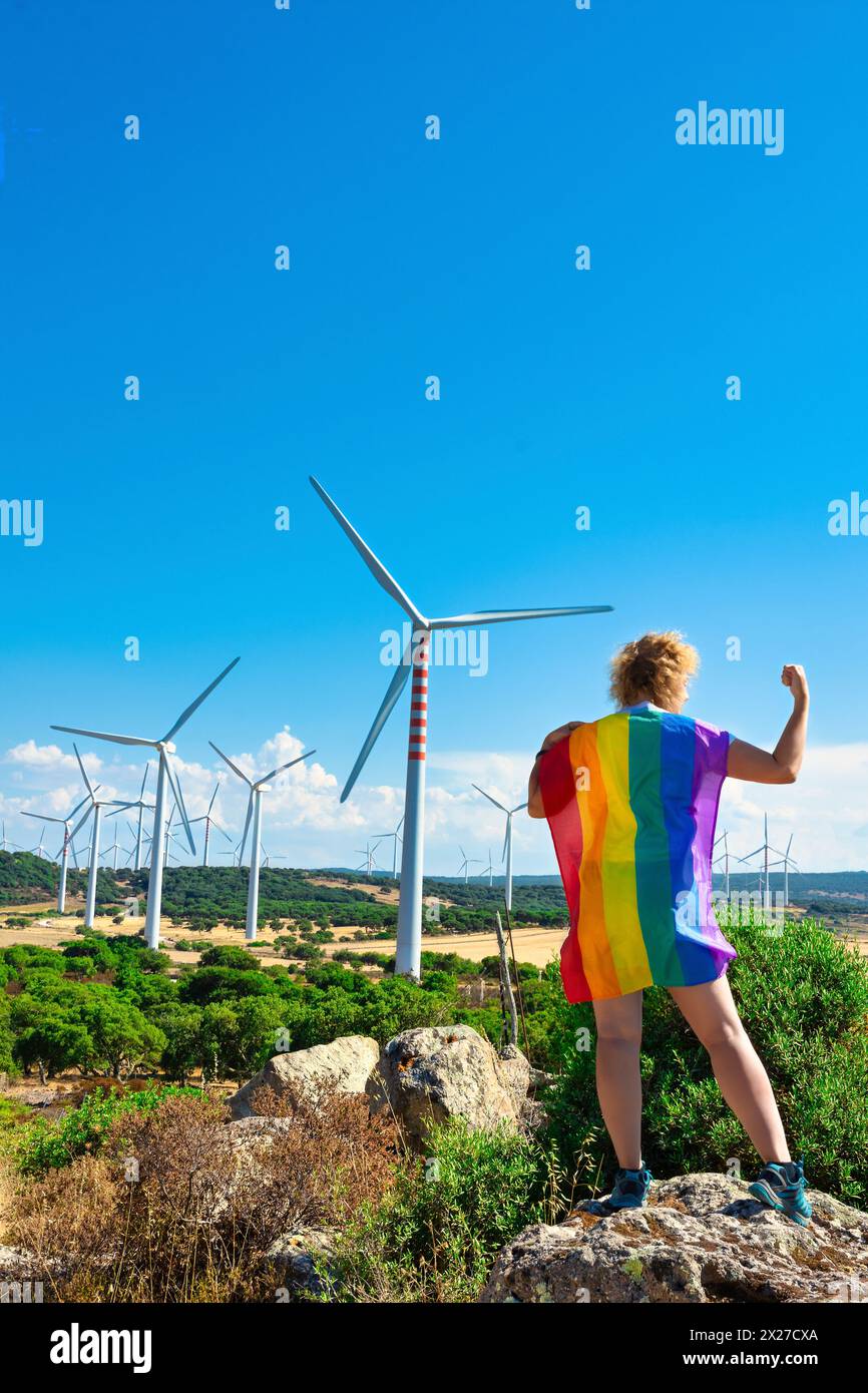 Donna adulta sulla schiena con una bandiera arcobaleno come mantello, su una roccia che piega il braccio e mostra il pugno come una forza in un paesaggio montuoso con il vento Foto Stock