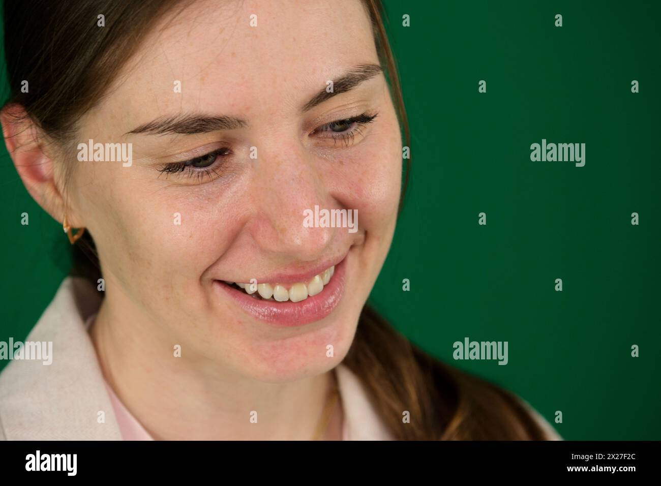 Giovane donna bianca bionda che ride mentre posa alla macchina fotografica in casa. su sfondo verde, cromakey, primo piano di una donna con emozioni in una giacca beige, manicure rossa, occhi blu Foto Stock