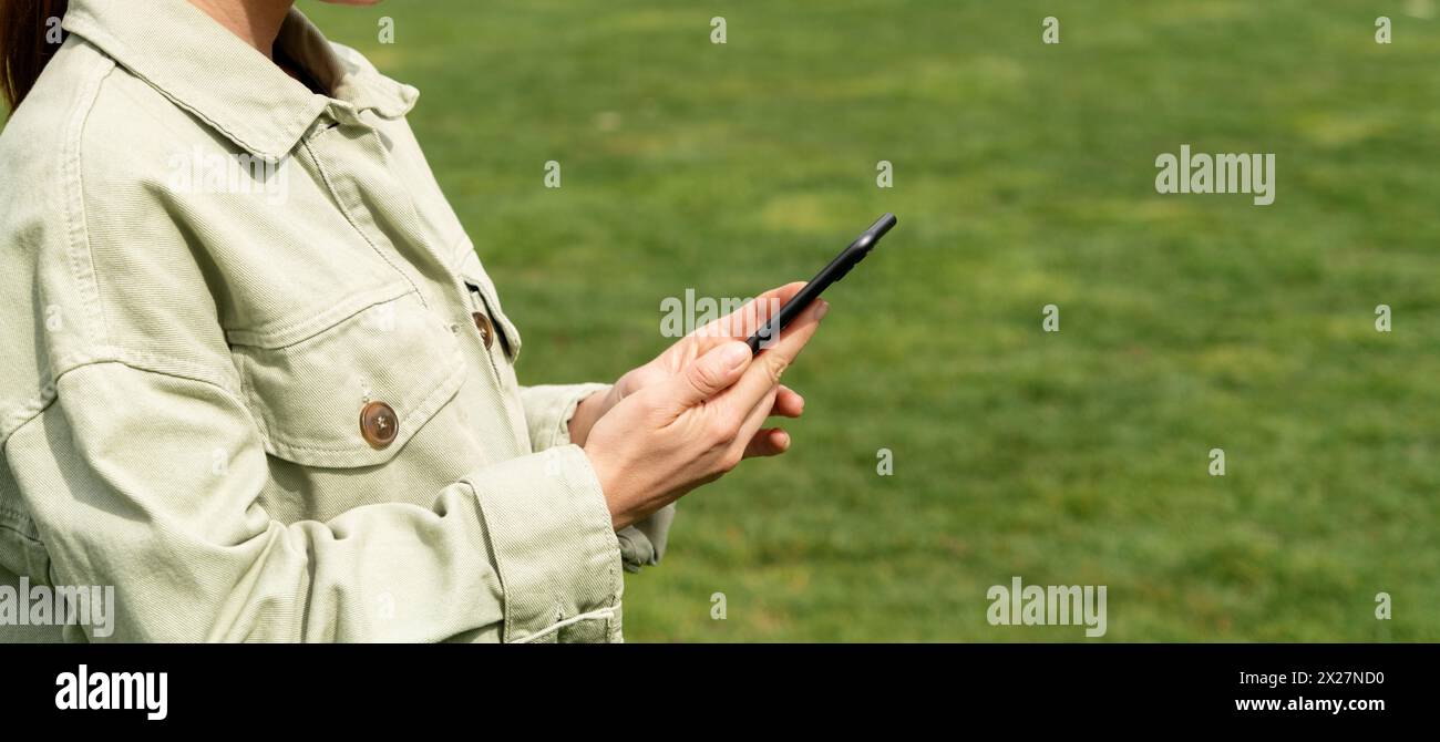 Una donna usa un telefono cellulare all'aperto sullo sfondo di un prato verde. Foto ravvicinata di smartphone nelle mani di una donna. Foto Stock