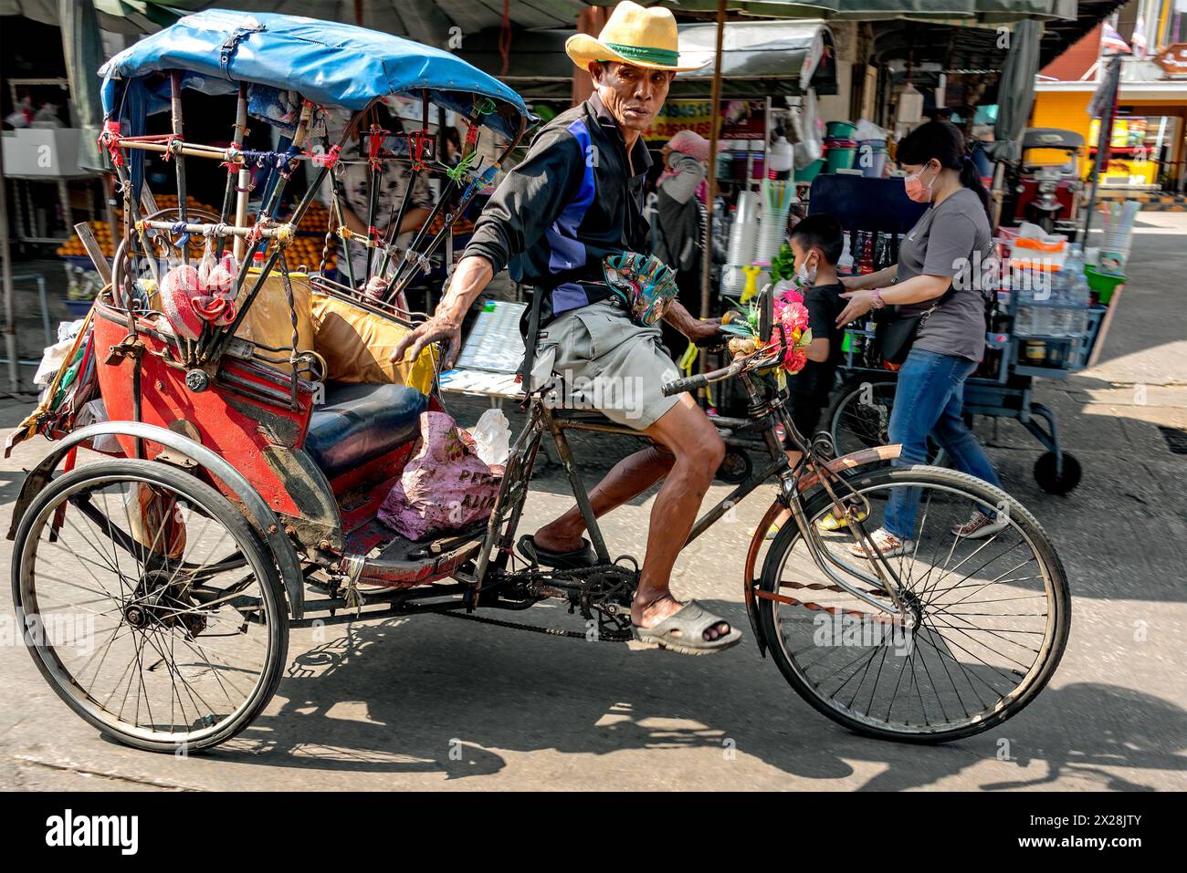 Samut Sakhon, Thailandia – 18 febbraio 2023: Di fronte alle bancarelle di frutta e bevande, un tassista triciclo dà un cartello prima di pedalare. Foto Stock