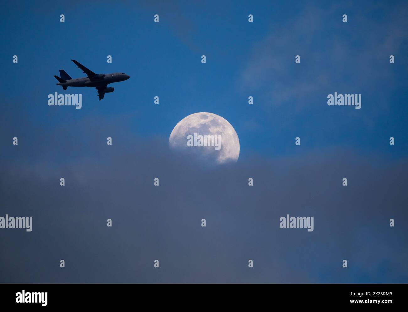 Un aereo di linea commerciale in avvicinamento finale a un aeroporto passa sopra una luna piena parzialmente oscurata da nuvole basse al tramonto. Foto Stock