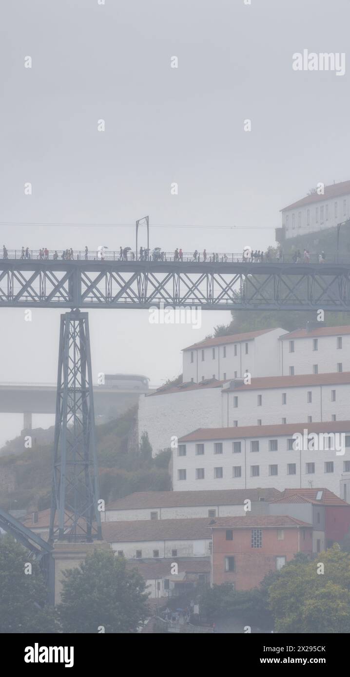 Turisti e pedoni che camminano con ombrelloni e impermeabili sulla piattaforma superiore del Ponte Dom Luis, circondati da nebbia e nuvole di pioggia bassa. Foto Stock