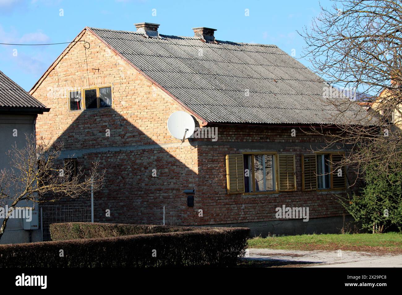 Piccola casa familiare urbana in mattoni rossi incompiuta con tende gialle a finestra in legno fatiscente e senza facciata accanto ad alti alberi senza foglie Foto Stock