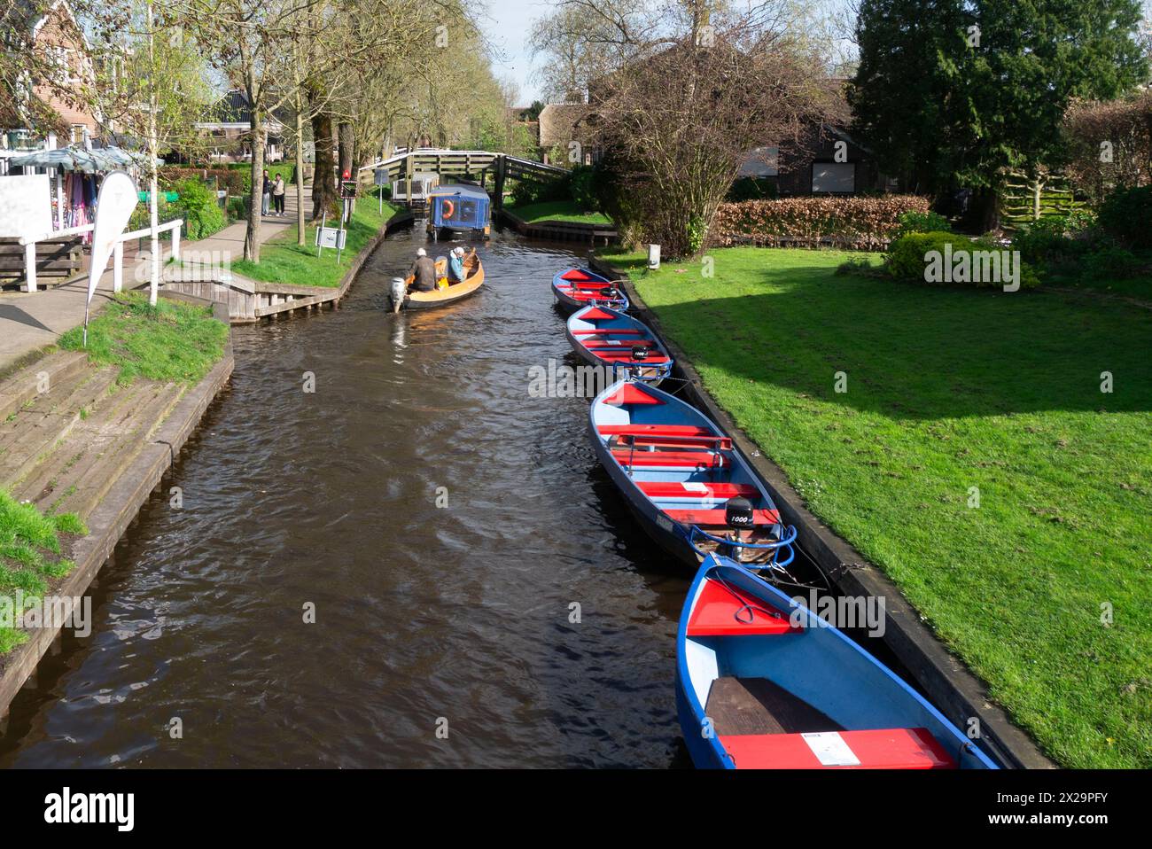 Due turisti che viaggiano lungo il canale in motoscafo con altre imbarcazioni a noleggio ormeggiate sul lato del villaggio del canale Giethoorn a Steenwijkerland, nella Venezia olandese Foto Stock