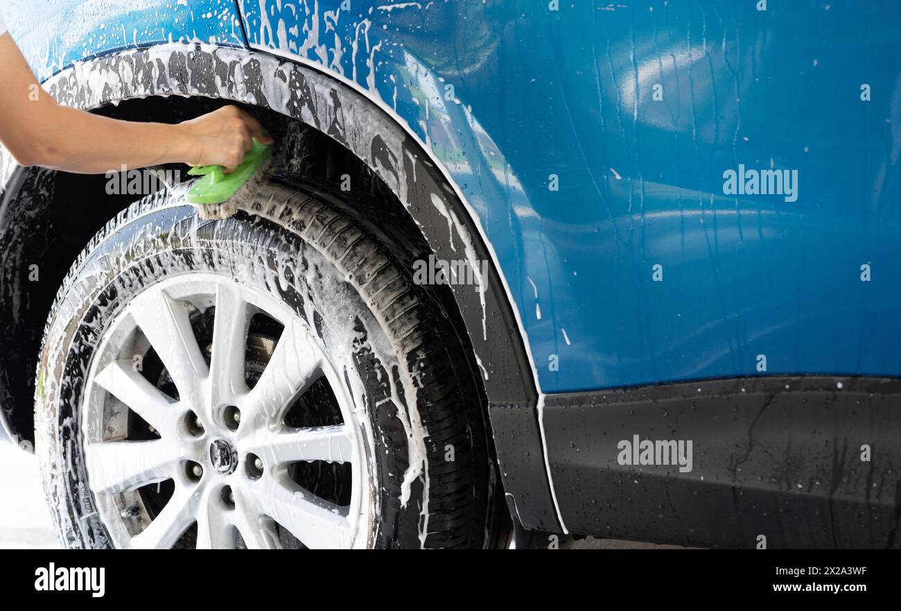 Una persona sta lavando una gomma di un'auto con un pennello verde. Lo pneumatico è coperto di acqua e sapone. Autolavaggio blu con schiuma di sapone bianca. Servizio Auto Care. Auto pulita Foto Stock