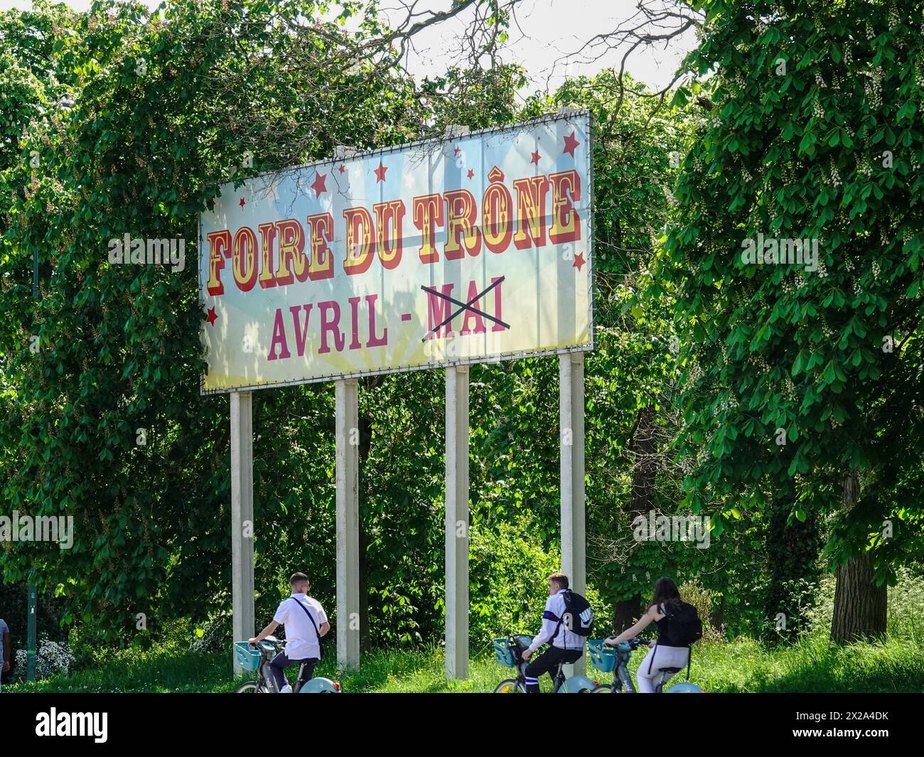 I giovani in bicicletta passano il cartello Foire du Trône, che pubblicizza il passaggio dal mese normale di maggio ad una data precedente di aprile a causa delle Olimpiadi. Foto Stock