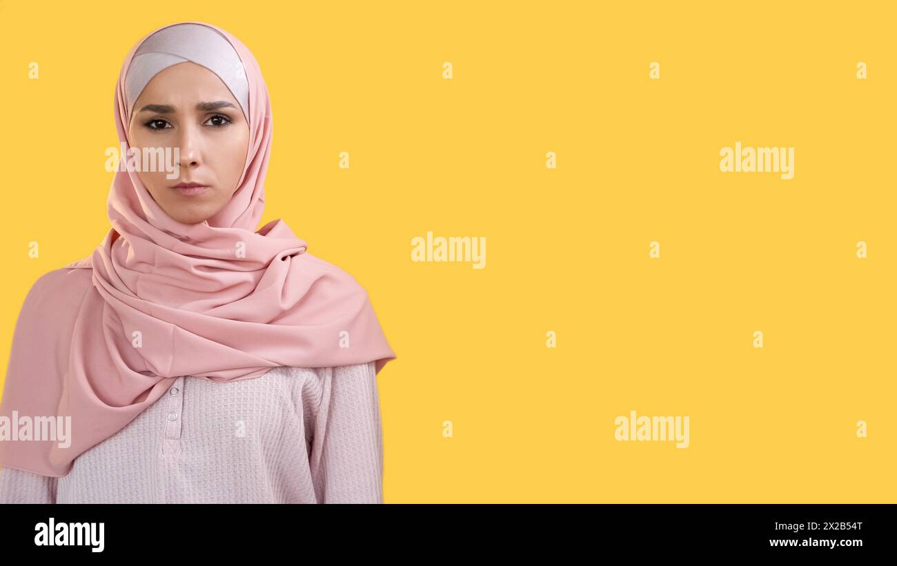 Ritratto femminile. Faccia seria. Donna sicura, severa arrabbiata e insoddisfatta in hijab che si aggira isolata su uno sfondo giallo vuoto. Foto Stock