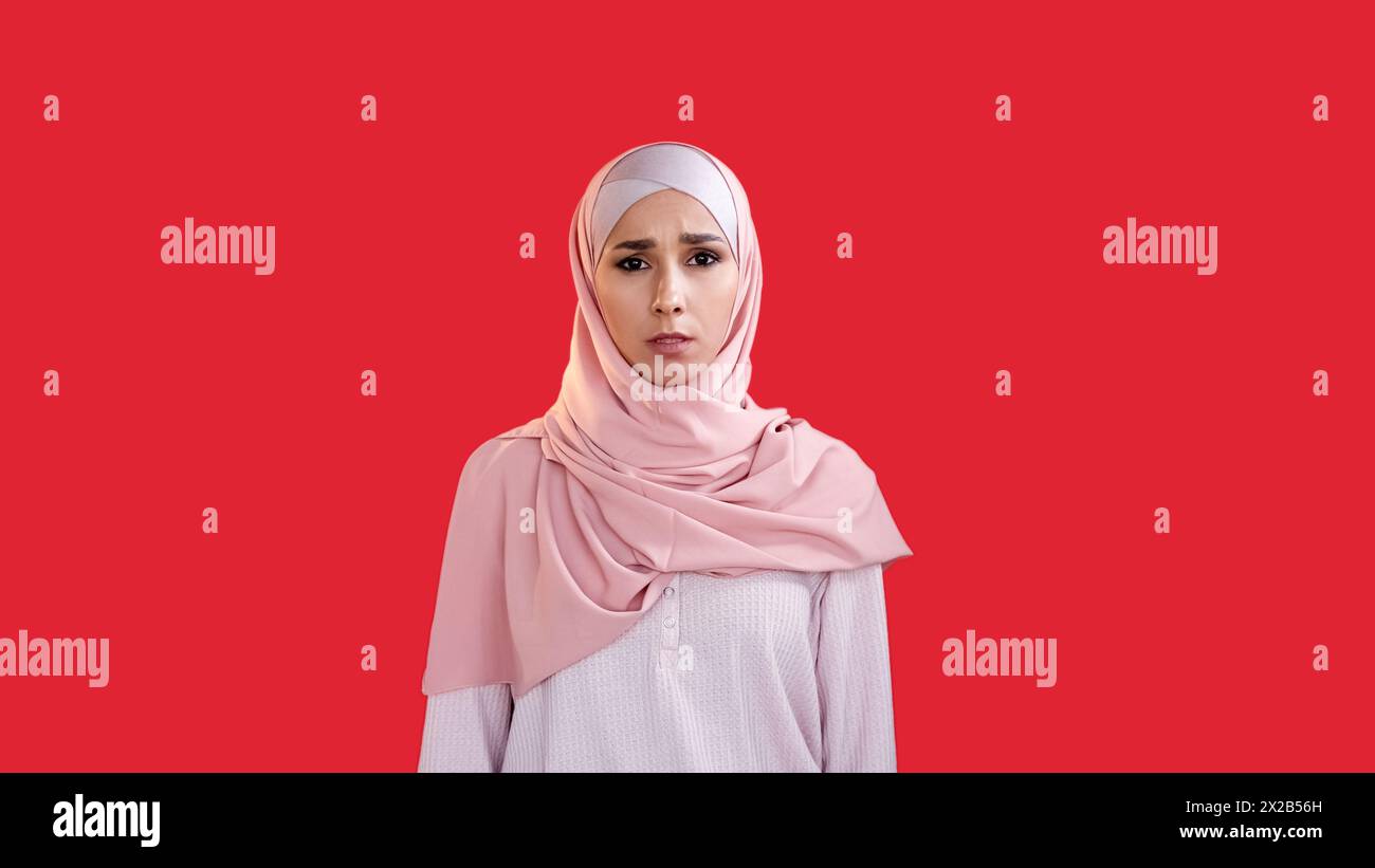 Donna disturbata. Faccia spaventata. Ansia da stress. Preoccupata ragazza spaventata in hijab con bocca aperta isolata su sfondo rosso vuoto. Foto Stock