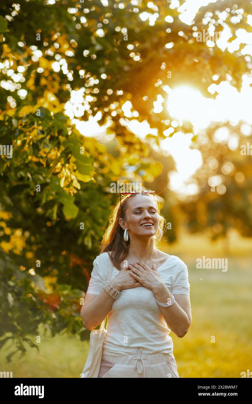 Ora legale. felice donna alla moda 40 anni in camicia bianca con mano sul petto nel parco cittadino. Foto Stock
