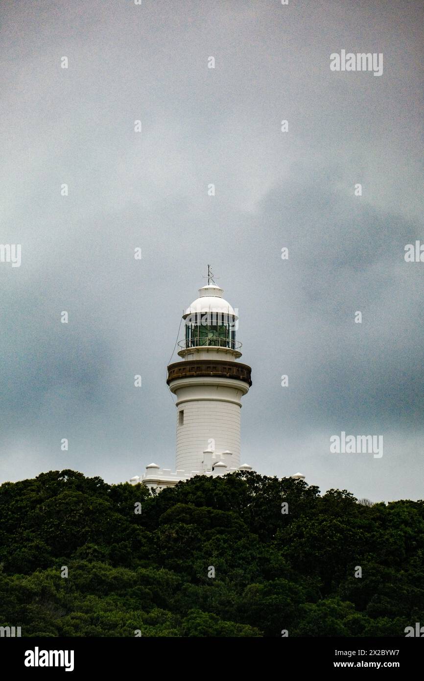 Arroccato in cima alle aspre scogliere, il faro di Byron Bay si erge alto come una sentinella costiera, il suo faro che penetra attraverso le nuvole in una giornata tranquilla Foto Stock