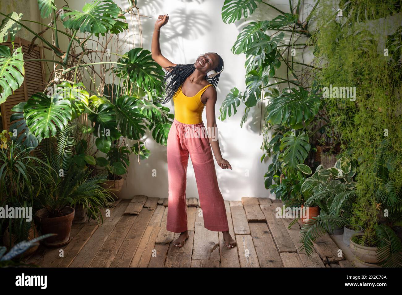 Felice donna nera spensierata che balla con gli occhi chiusi ascoltando musica in cuffie tra le piante d'casa Foto Stock