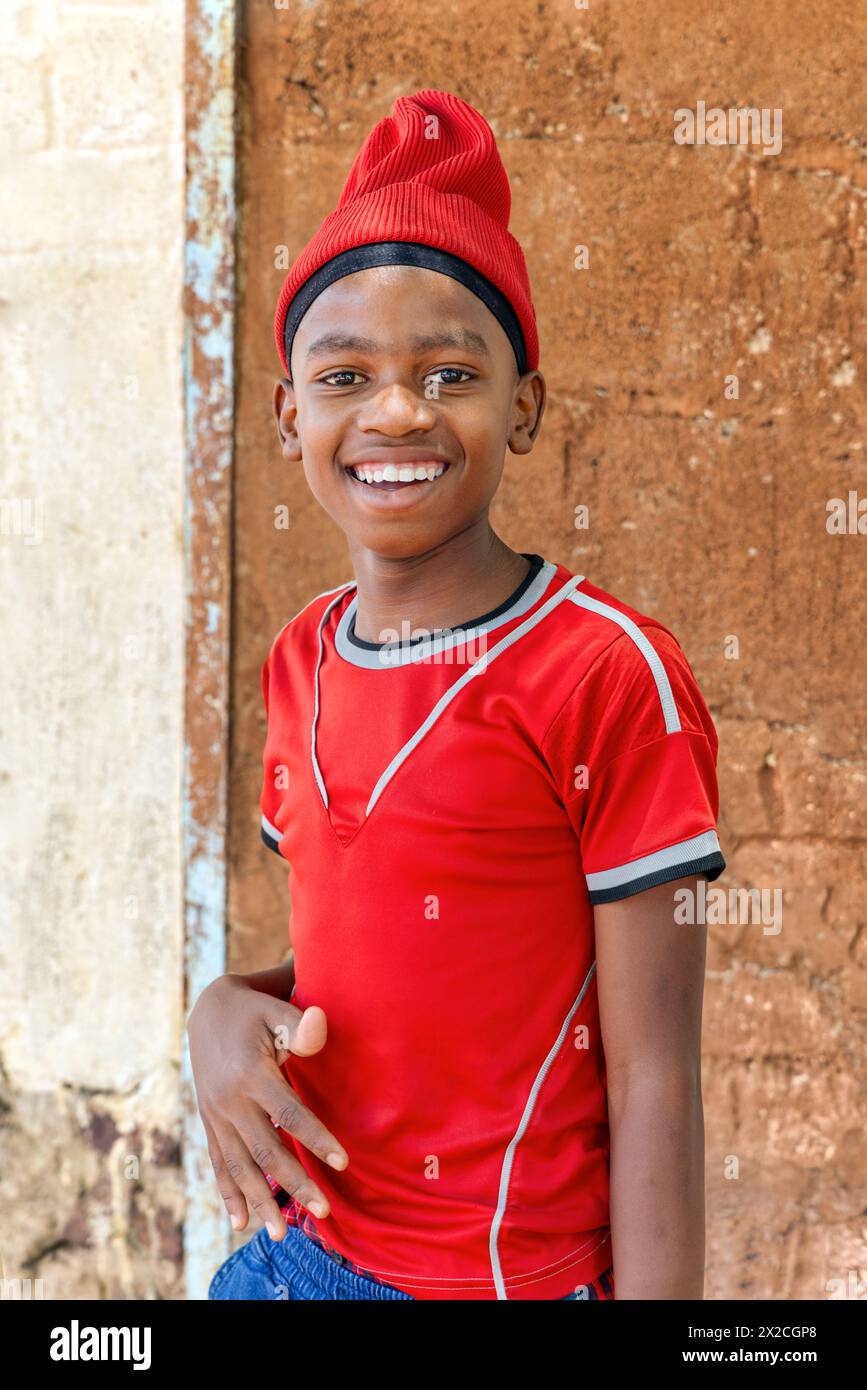 sorridente bambino africano del villaggio con un berretto che gioca all'aperto di fronte alla casa in un villaggio, Foto Stock