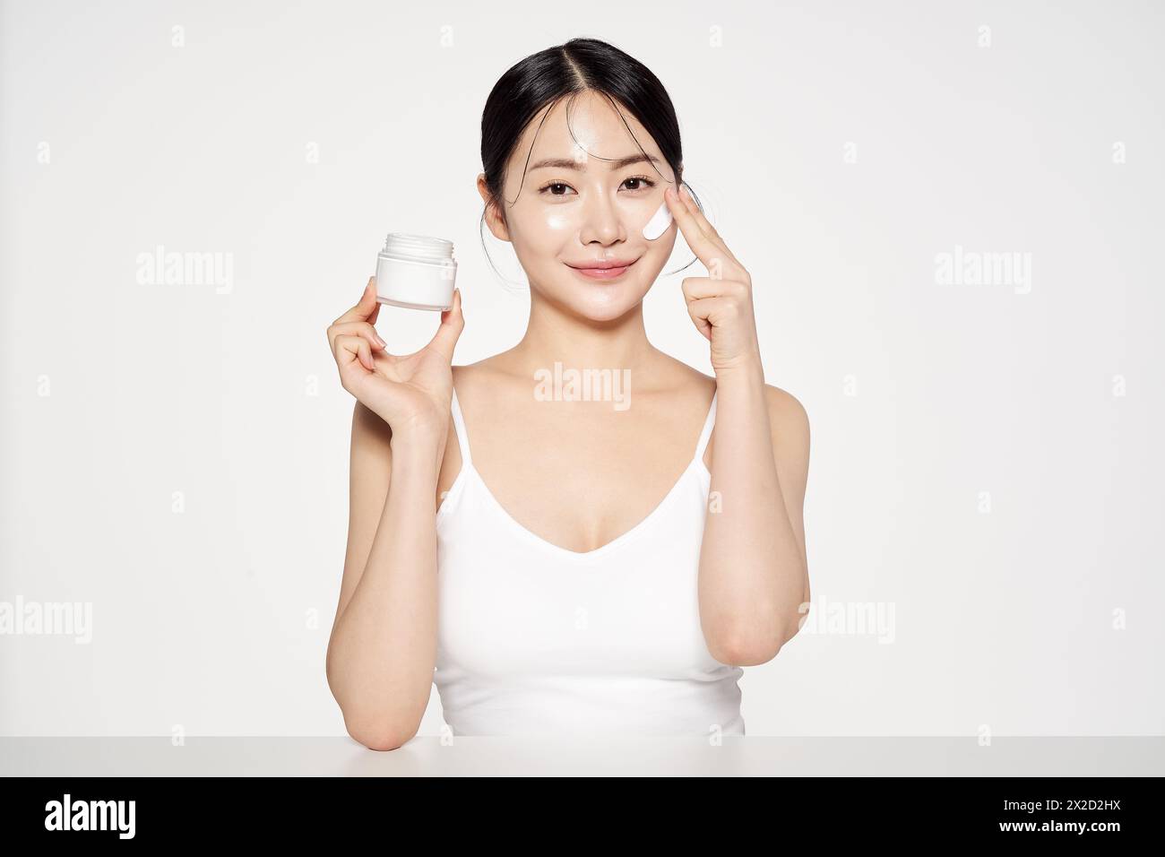 Una donna asiatica che tiene un contenitore di crema e le applica cosmetici sul viso Foto Stock