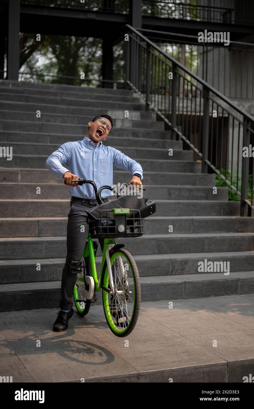 Un esuberante uomo d'affari millenario asiatico cavalca con gioia una bicicletta verde giù per le scale, esprimendo felicità e libertà. concetto di uomini d'affari Foto Stock