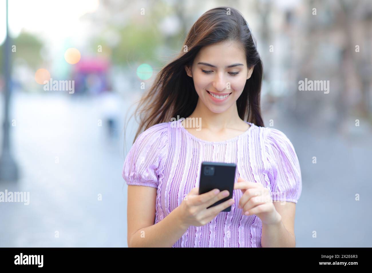 Vista frontale ritratto di una donna felice che cammina usando il cellulare per strada Foto Stock