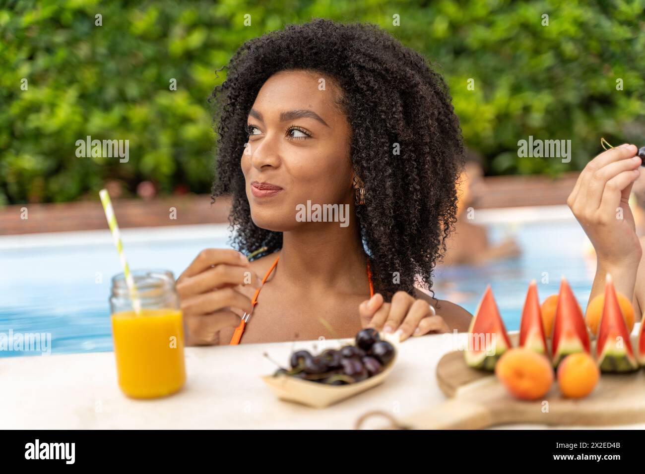 Giovane donna che sorseggia succhi di frutta a bordo piscina - gusta spuntini salutari - ambiente estivo rilassato, rinfresco naturale. Foto Stock
