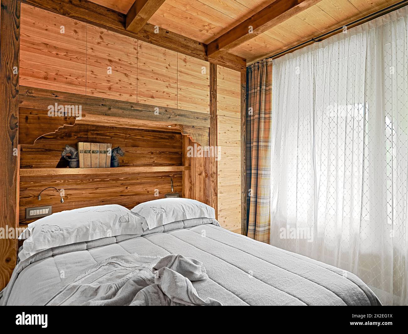 interno rustico della camera da letto con mobili in legno e soffitto in legno sulla destra la grande finestra con tenda Foto Stock
