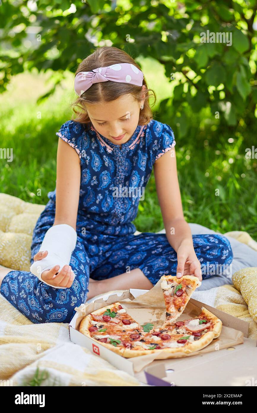 Una ragazza con un abito blu e un cast mangia la pizza all'aperto in una giornata di sole. Foto Stock