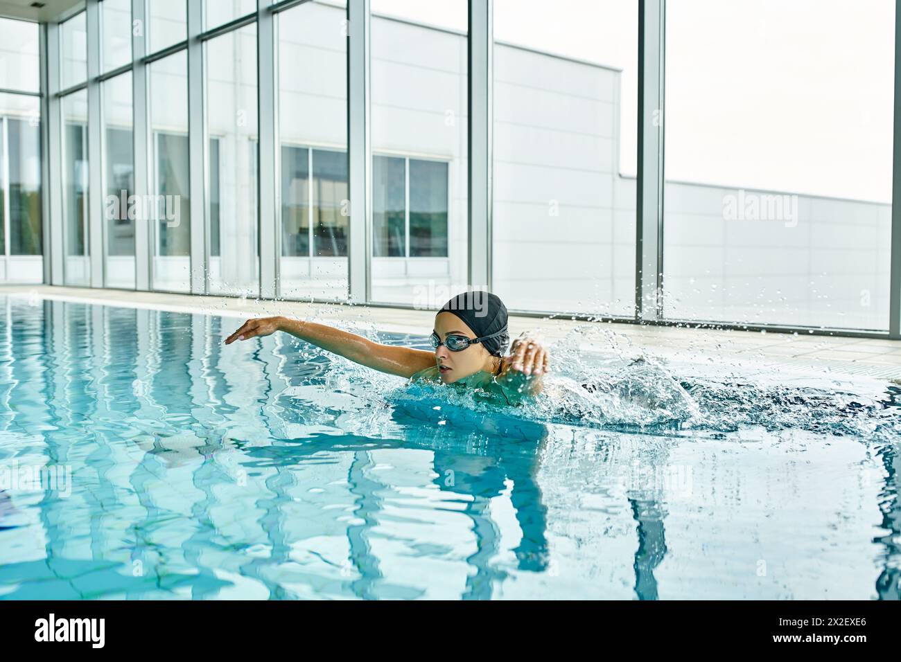 Una giovane donna con costume da bagno e cuffia che nuota graziosamente in una piscina interna all'interno di un edificio. Foto Stock