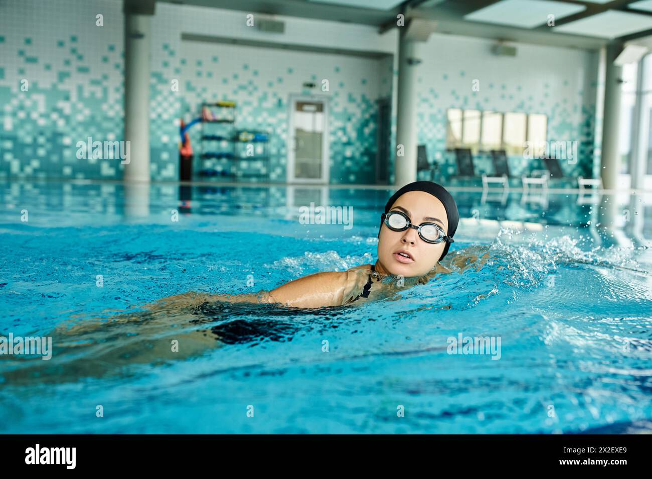 Una giovane donna che indossa occhiali di protezione, un costume da bagno e una cuffia che nuota con grazia in una piscina coperta. Foto Stock