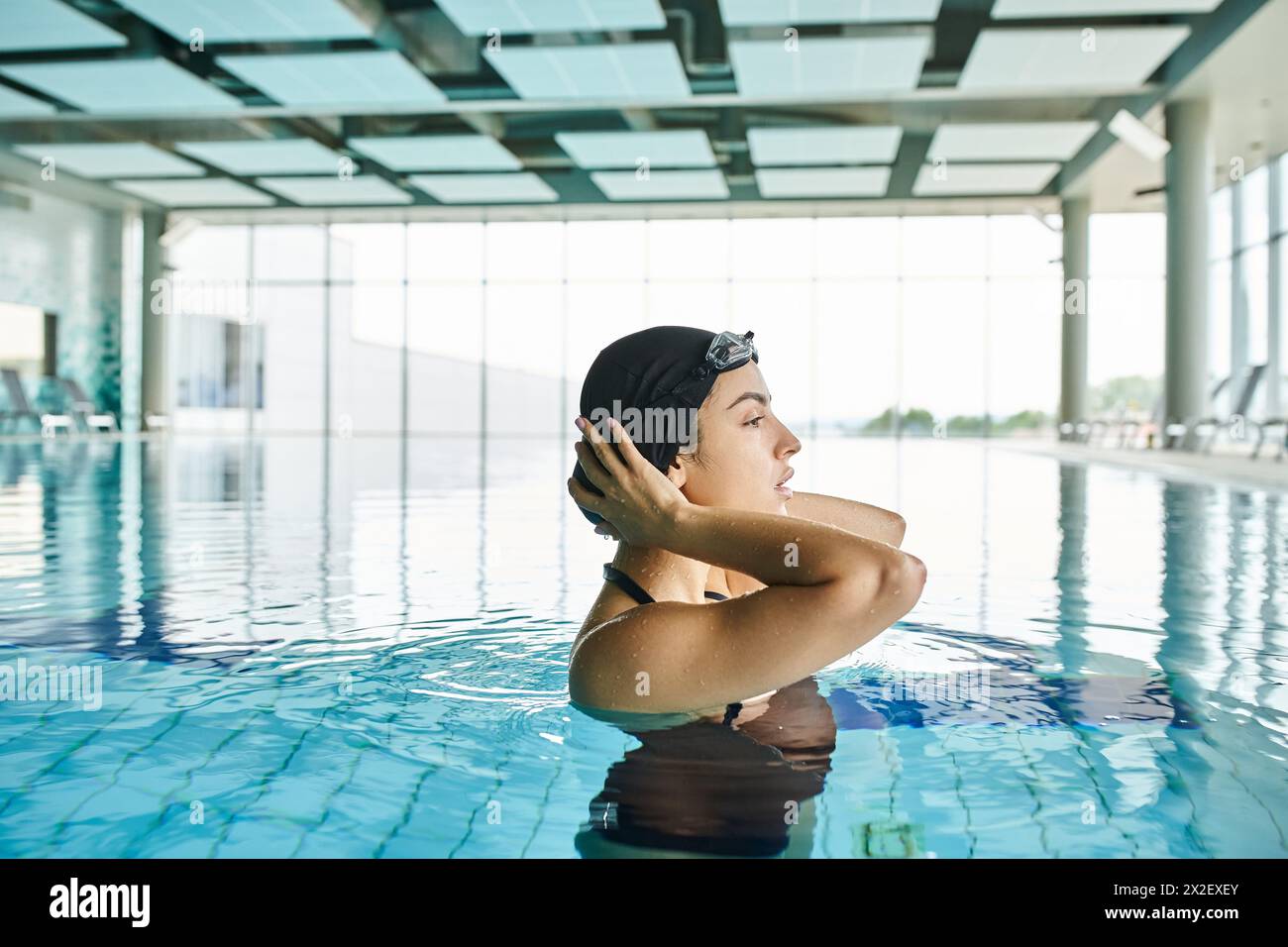 Una giovane donna con costume da bagno e cuffia nuota con grazia in una spa interna, indossando occhiali di protezione. Foto Stock