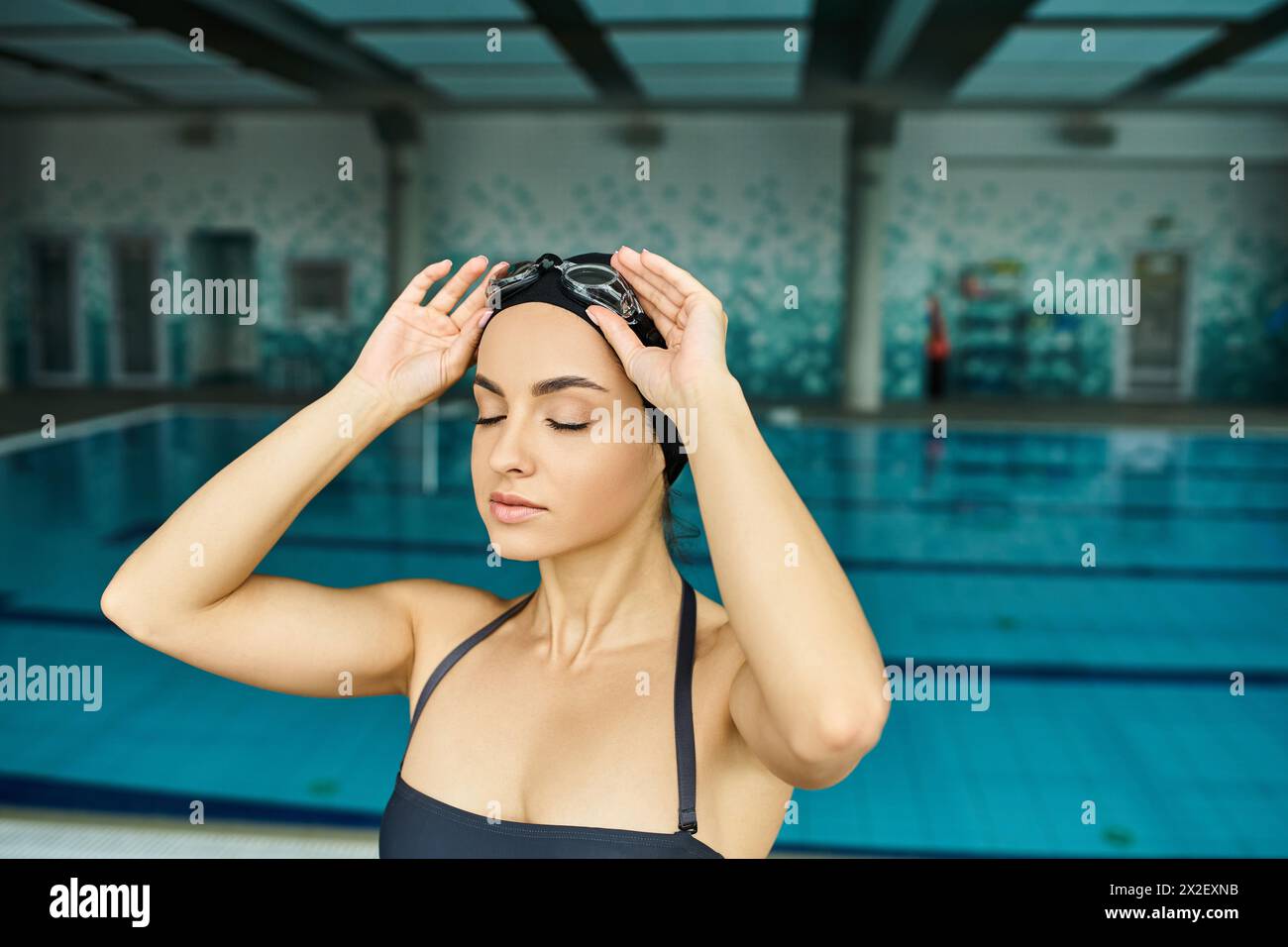 Una giovane donna con costume da bagno e cuffia che tiene la testa davanti a una piscina coperta. Foto Stock